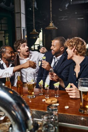 cheerful interracial friends holding tequila shots with lime near glasses of beer on bar counter