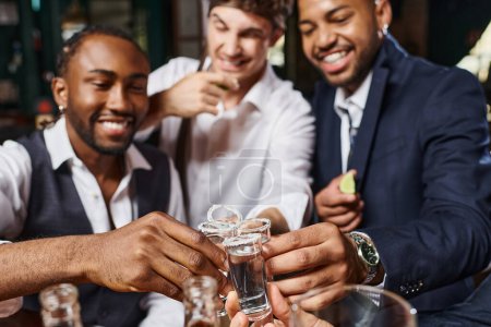 focus of tequila shots, cheerful interracial friends toasting with glasses during bachelor party