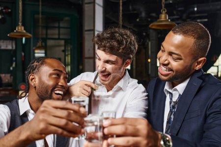 happy interracial friends toasting with glasses of tequila shots and laughing during bachelor party