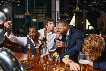 candid photo of funny and drunk multicultural men in formal wear drinking tequila in bar after work