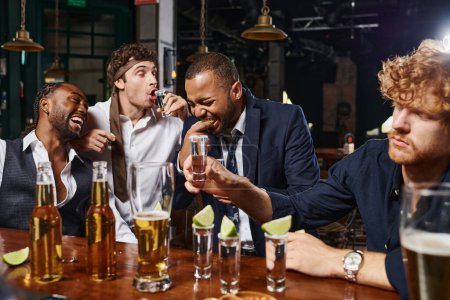 candid photo of funny and drunk interracial men in formal wear drinking tequila in bar after work