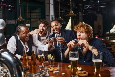 Gruppe von vier fröhlichen und betrunkenen interrassischen Männern in offizieller Kleidung trinken Tequila in der Bar nach der Arbeit