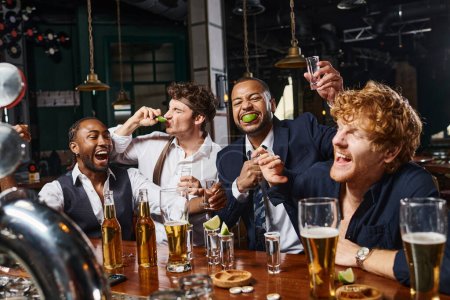 Gruppe von vier fröhlichen und betrunkenen multiethnischen Freunden in formeller Kleidung trinken Tequila in der Bar nach der Arbeit