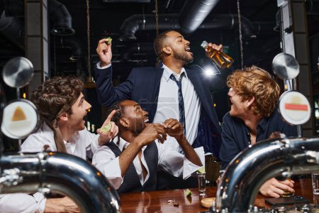 hombres felices mirando a un amigo afroamericano bebiendo cerveza de dos botellas después del trabajo en el bar