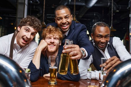 group of happy and drunk multiethnic friends in formal wear holding tequila shot and beer in bar