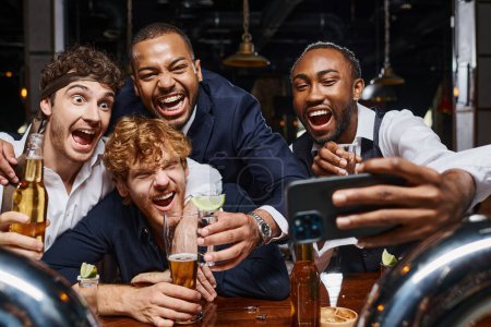 Aufgeregte multiethnische Kollegen in offizieller Kleidung machen nach Feierabend ein Selfie mit dem Smartphone in der Bar