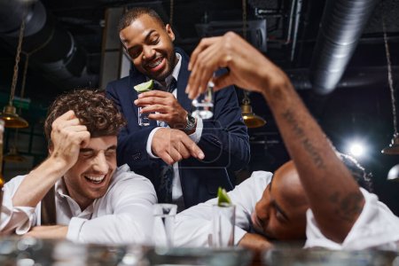three drunk interracial friends drinking tequila shots and laughing while relaxing after work in bar
