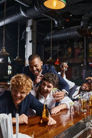 drunk and funny interracial men in formal wear laughing while drinking tequila and beer in bar