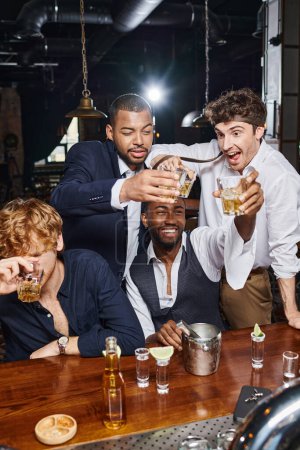 drunk and funny interracial men toasting with glasses of whiskey near beer and tequila shots in bar