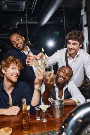 vier fröhliche multikulturelle Freunde stoßen mit Gläsern Whiskey in der Nähe von Tequila-Shots und Bier in der Bar an