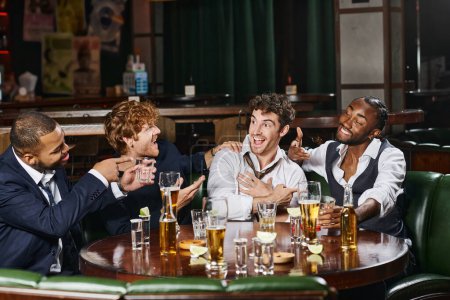happy multiethnic men pointing at friend in bar, male friends during bachelor party in bar