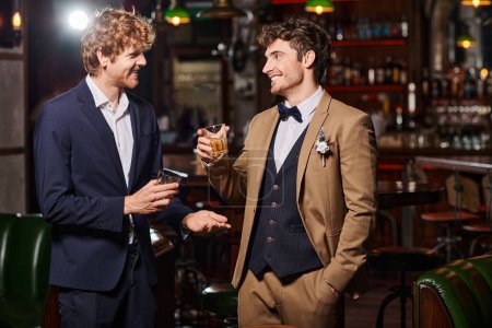Téléchargez les photos : Marié heureux en costume avec boutonnière et témoin tenant des verres de whisky pendant la fête de célibataire - en image libre de droit