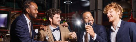 happy interracial best men in suits holding glasses of whiskey and congratulating friend, banner
