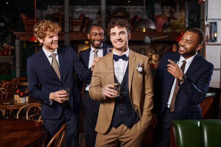 Junggesellenabschied, glückliche exotische Trauzeugen, die den Bräutigam im Anzug mit Whiskey in der Bar anschauen