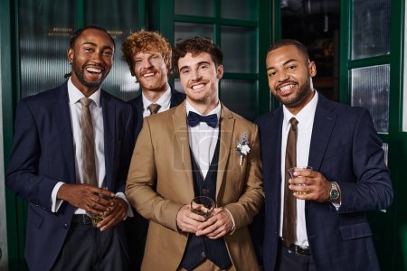 Téléchargez les photos : Enterrement de vie de garçon, portrait de meilleurs hommes et mariés multiethniques debout avec des verres de whisky - en image libre de droit