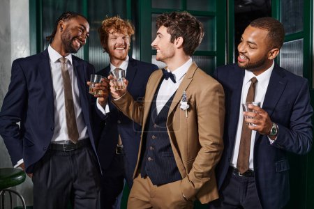 Foto de Despedida de soltero, multiétnicos mejores hombres tostando vasos de whisky con elegante novio en el bar - Imagen libre de derechos