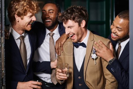 Junggesellenabschied, aufgeregte interrassische Trauzeugen und Bräutigam lachen und halten Gläser Whiskey