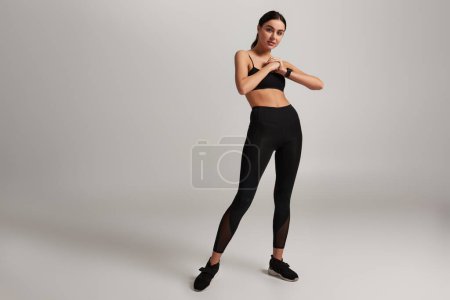 Foto de Joven mujer deportiva en ropa deportiva negra con rastreador de fitness en la muñeca haciendo ejercicio sobre fondo gris - Imagen libre de derechos