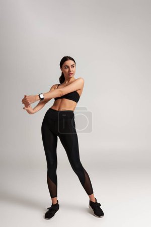 hübsche sportliche Frau in schwarzer Sportbekleidung mit Fitness-Tracker am Handgelenk, die sich vor grauem Hintergrund dehnt