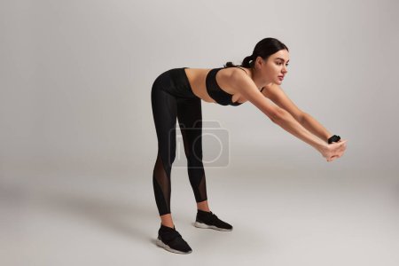 brunette woman in black sportswear with fitness tracker on wrist stretching back on grey backdrop