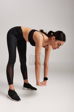 Foto de Mujer motivada en el desgaste activo negro con rastreador de fitness en la muñeca que se extiende hacia atrás en el fondo gris - Imagen libre de derechos