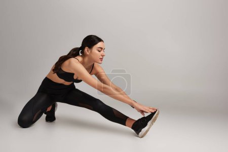 Foto de Atractiva mujer en desgaste activo negro con rastreador de fitness en la pierna de estiramiento de la muñeca sobre fondo gris - Imagen libre de derechos