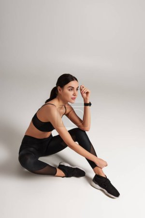 Nachdenkliche Sportlerin in schwarzer Aktivkleidung mit Fitness-Tracker am Handgelenk sitzt auf grauem Hintergrund