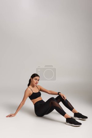 müde Sportlerin in schwarzer Aktivkleidung mit Fitness-Tracker am Handgelenk auf grauem Hintergrund