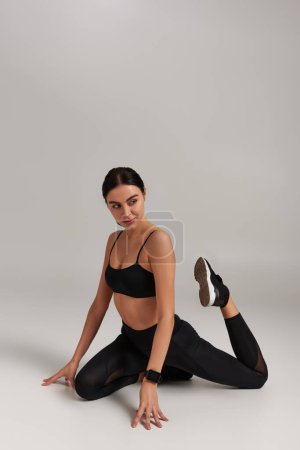 Foto de Joven deportista flexible en leggins negros y piernas de estiramiento de la parte superior de la cosecha en el fondo gris - Imagen libre de derechos