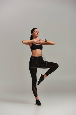 femme calme en tenue active méditant et debout sur une jambe sur fond gris, technique de respiration