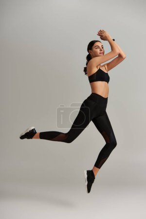 junge, fitte Frau in schwarzen Leggings, bauchfreiem Oberteil und Turnschuhen, die auf grauem Hintergrund springen, schweben