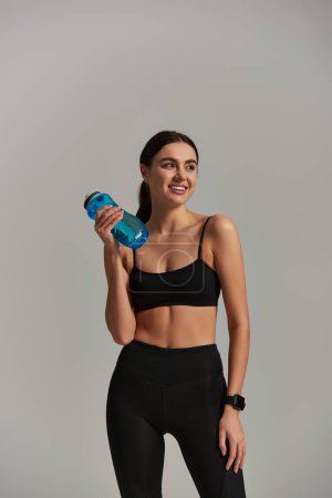 happy slim sportswoman in leggings and crop top holding bottle with water on grey background