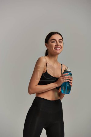 cheerful slim sportswoman in leggings and crop top holding bottle with water on grey background
