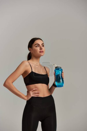 slim sportswoman in leggings and crop top holding bottle with water while standing with hands on hip