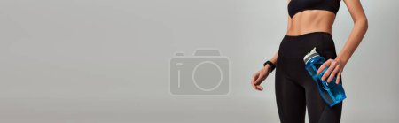 cropped woman in leggings and crop top holding bottle with water on grey background, banner