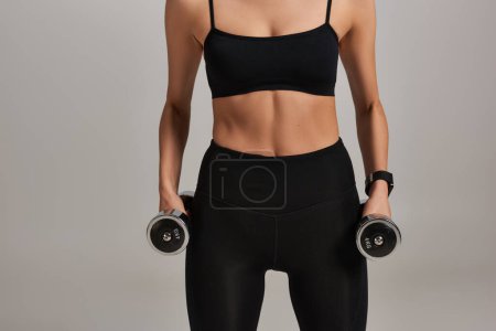 Foto de Vista recortada de una deportista fuerte en desgaste activo negro haciendo ejercicio con pesas sobre fondo gris - Imagen libre de derechos