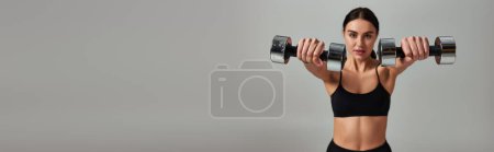motivated sportswoman in black active wear working out with dumbbells on grey backdrop, banner Poster 677588414