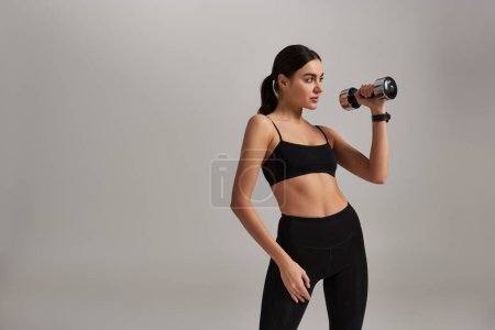 slim and strong young woman in active wear lifting dumbbell while working out on grey backdrop
