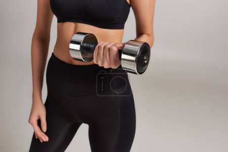 cropped shot of slim sportswoman in active wear lifting dumbbell while working out on grey backdrop