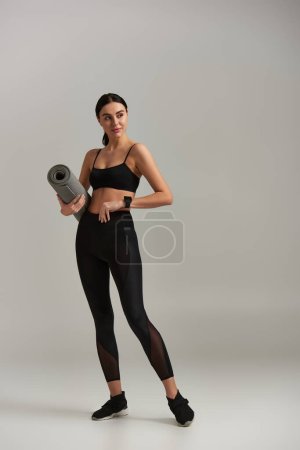 full length of fit sportswoman in active wear holding fitness mat and standing on grey background