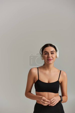 attractive sportswoman in crop top listening music in wireless headphones on grey background