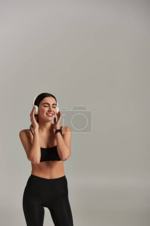 deportista alegre en ropa activa escuchando música en auriculares inalámbricos sobre fondo gris