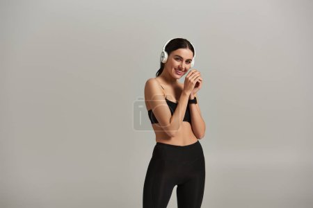 zufriedene Sportlerin in aktiver Kleidung hört Musik in drahtlosen Kopfhörern auf grauem Hintergrund