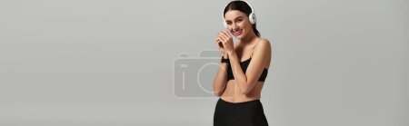 pleased sportswoman in active wear listening music in wireless headphones on grey background, banner