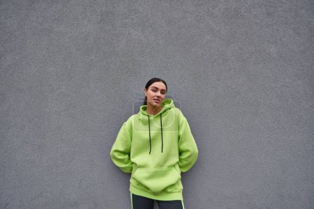 beautiful sportswoman in trendy lime color hoodie standing near grey concrete wall outdoors