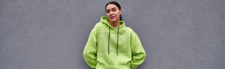 beautiful sportswoman in trendy lime color hoodie standing near grey concrete wall outdoors, banner