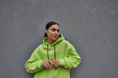 brunette young woman in trendy lime color hoodie looking away near grey concrete wall outdoors