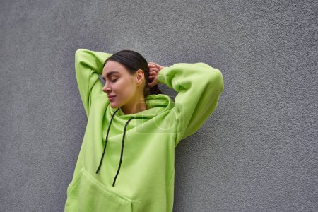pretty young woman in trendy lime color hoodie posing while standing near grey concrete wall