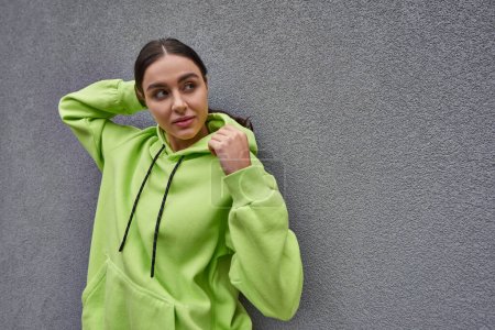 brunette young woman in trendy lime color hoodie posing while standing near grey concrete wall