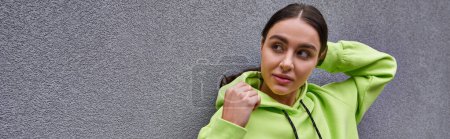 morena mujer joven en moda color lima sudadera con capucha posando mientras está de pie cerca de la pared de hormigón, pancarta